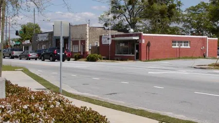 Small town street with brick buildings, cars, trees, and a sidewalk on a sunny day with blue sky.