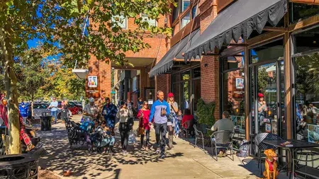 People walking in Woodstock GA and sitting outside cafes along a sunny urban sidewalk with trees and brick buildings.