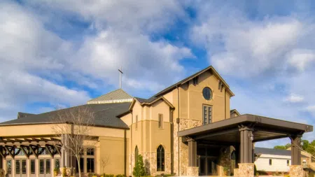 Modern church building with stone and beige walls under a partly cloudy blue sky and a cross on the roof.