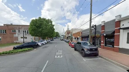 Downtown street view with parked cars, shade trees, brick buildings, and a partly cloudy sky overhead.