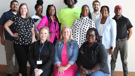 Diverse group of smiling professionals posing indoors against a plain white wall in casual and business attire.