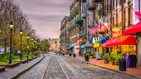 Cobblestone street with tram tracks lined by colorful shops, flags, and street lamps at sunset.