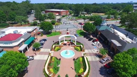 Aerial view of a town square with a central fountain, landscaped gardens, and surrounding shops under clear sky.