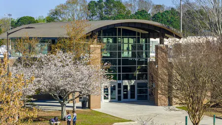 Modern building with large glass windows surrounded by blooming trees and people walking on a sunny day.