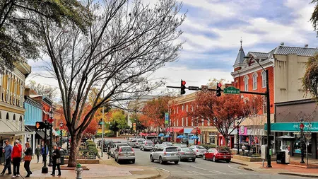 Busy small-town street with colorful buildings, autumn trees, vehicles, and pedestrians under a partly cloudy sky.