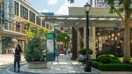 Urban shopping area with pedestrians, modern buildings, greenery, benches, and street lamps on a sunny day