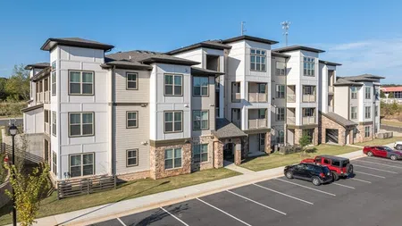 Modern three-story apartment complex with stone and white siding exterior and a parking lot with several cars.