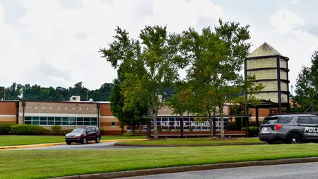Exterior view of a school building with green trees, lawn, and police vehicle parked outside on a cloudy day
