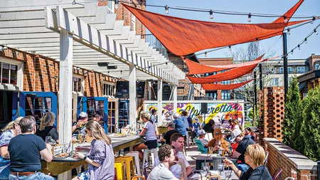 Outdoor restaurant patio with people dining under red sun shades and string lights on a sunny day