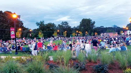 Large crowd gathered in a park during evening event with street lamps and trees under a cloudy sky