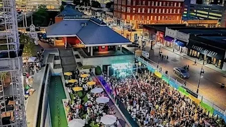 Crowd gathered at an outdoor event in an urban plaza with seating, umbrellas, and city buildings at dusk.