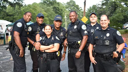 Group of seven smiling police officers standing together outdoors in uniform on a street with trees in the background