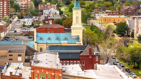 Cityscape with historic church steeple, colorful buildings, and green trees under a partly cloudy blue sky.