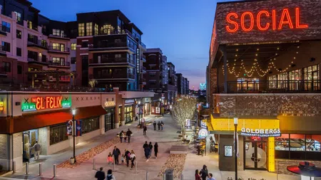 Evening view of a lively urban pedestrian street with restaurants, neon signs, and modern buildings under blue sky.