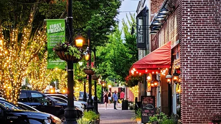 Downtown street decorated with string lights, brick buildings, parked cars, and pedestrians strolling at dusk
