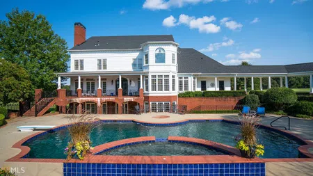 Large white two-story house with a wraparound porch and curved swimming pool under a blue sky.