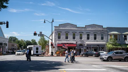 People crossing a sunny street at an intersection with historic two-story buildings and shops in the background