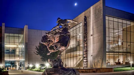 Bronze cowboy statue on horseback at night in front of a modern museum building under a crescent moon