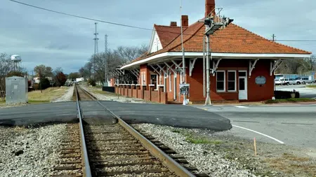 Historic red brick train station near railroad tracks under a cloudy sky in a small town setting.
