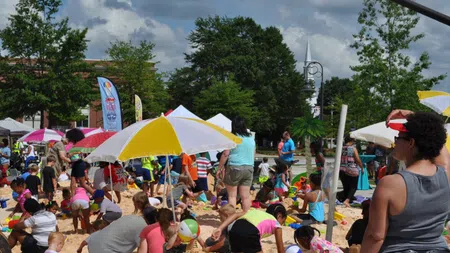 Children and adults enjoying a sunny day at a crowded outdoor sand play area with colorful umbrellas and toys.