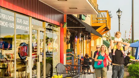 Families and people walking along a sunny small-town street with shops, outdoor seating, and street lamps.