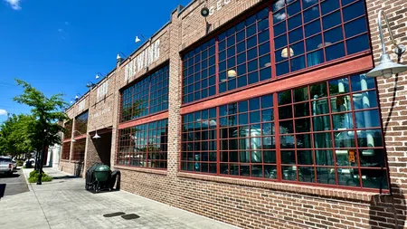 Brick building with large red-framed windows under blue sky on a sunny day in an urban setting
