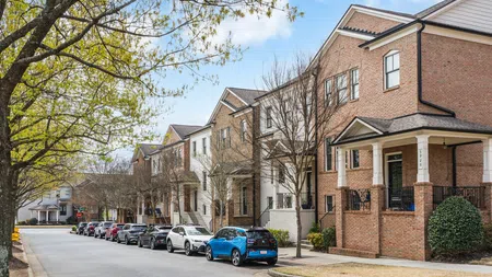 Residential street lined with modern brick townhouses and parked cars under a partly cloudy sky in spring.