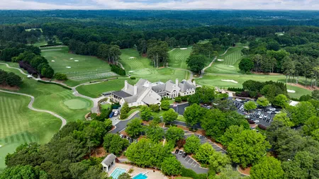 Aerial view of a golf course with clubhouse, parking, swimming pool, and surrounding trees under blue sky.
