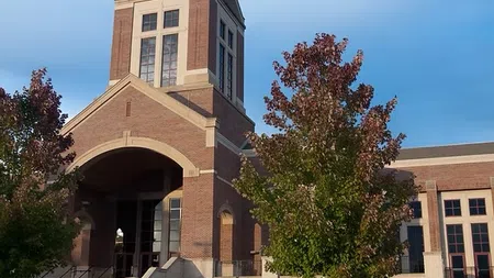 Brick church building with tall bell tower and cross under clear blue sky with trees in front.
