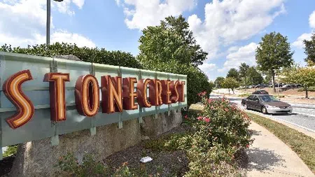 Stonecrest sign with large orange letters beside a landscaped sidewalk and a car on a sunny day with blue sky.