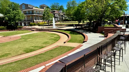 Outdoor plaza with curved brick pathways, metal chairs, water fountain, green lawn, and surrounding buildings under a clear blue sky