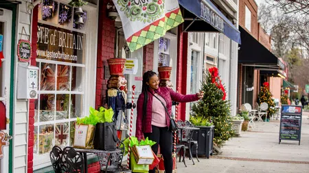 Woman taking selfie by Christmas-decorated shops with shopping bags and festive nutcracker statues on sidewalk.