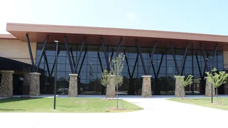 Modern building with large glass windows and stone pillars under a brown angled roof on a grassy lawn.