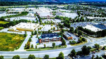 Aerial view of a large shopping plaza with multiple stores, extensive parking lots, and surrounding greenery under a clear sky.
