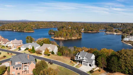 Aerial view of lakeside neighborhood with houses, trees, and a large lake under blue sky with scattered clouds.