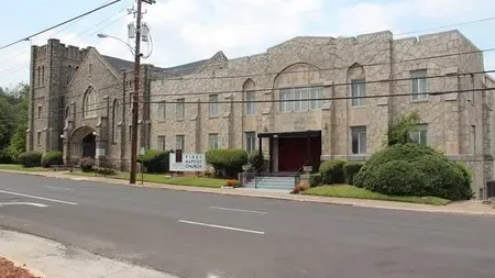 Stone facade of First Baptist Church with arched windows and trimmed bushes along the roadway