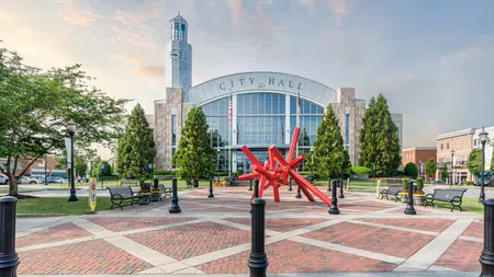 Modern city hall building with a tall clock tower, red abstract sculpture, and landscaped plaza at sunset.