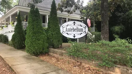 Historic Antebellum house with white porch, lush greenery, and decorative sign along a stone-bordered sidewalk.