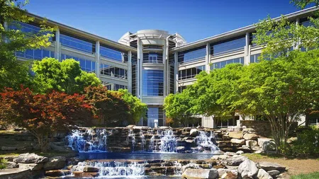 Modern office building with glass facade behind landscaped waterfall and lush green and red trees under blue sky
