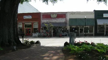 Small town street with vintage shops, awnings, outdoor seating, and large tree shading a brick sidewalk.
