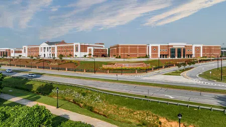 Wide view of a large brick educational campus with multiple buildings under a partly cloudy sky.