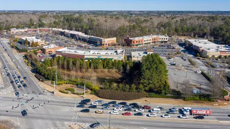 Aerial view of suburban shopping center with parked cars, adjacent forest, and busy multi-lane road with vehicles.