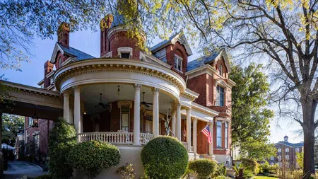Historic red brick Victorian house with round porch, white columns, lush greenery, and American flag on a sunny day.