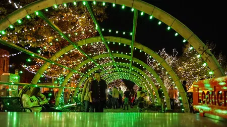 People walk and sit under vibrant green arched lights with illuminated trees at night in a festive outdoor setting