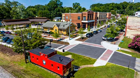 Small town intersection with vintage red train caboose, modern buildings, trees, and parked cars on a sunny day.
