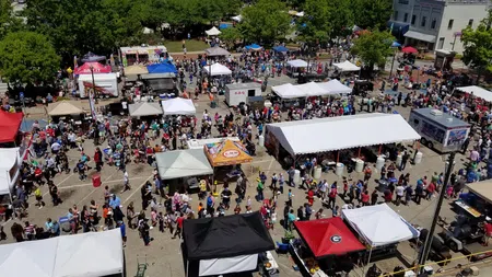Overhead view of a busy outdoor festival with food tents, crowds, and surrounding trees and buildings on a sunny day