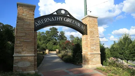 Stone pillars with a black arch reading Abernathy Greenway over a pathway surrounded by trees and blue sky.