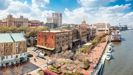 Historic riverside district with brick buildings, tree-lined walkway, boats docked along the river under a blue sky.