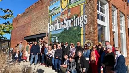 Group of people posing in front of a Powder Springs welcome mural on a sunny day.
