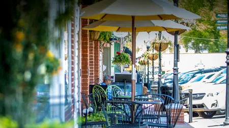 Outdoor café seating with umbrellas along a sunny street lined with parked white cars and hanging flower baskets.